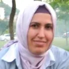 A close-up portrait of a smiling woman wearing a light-colored headscarf and shirt outdoors.