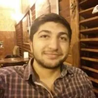 İrfan, ahşap panellerle kaplı sıcak aydınlatmalı bir mekanda hafif gülümseyerek kameraya bakıyor.