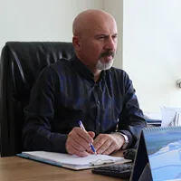 A man in a dark shirt sitting at an office desk and writing on paper with a blue pen.