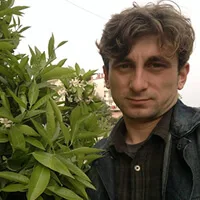 A man with dark hair and a jacket stands next to a green-leafed plant with small white flowers.