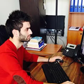 A bearded man wearing a red sweater works at a computer in an office.