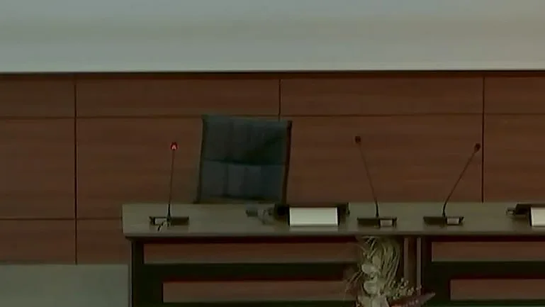Konferans salonunda kürsüde konuşan bir kişi ve yanındaki boş panel masası.