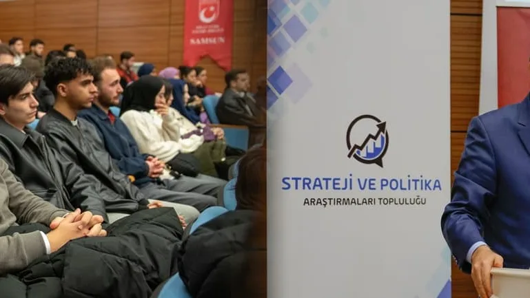 Salonda oturan dinleyiciler ve “Strateji ve Politika Araştırmaları Topluluğu” pankartının yanındaki kürsüde konuşan bir kişi.