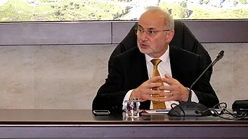 Two people talk facing each other from either end of a meeting table, a Turkish flag on the left and a mountain landscape on the wall behind.