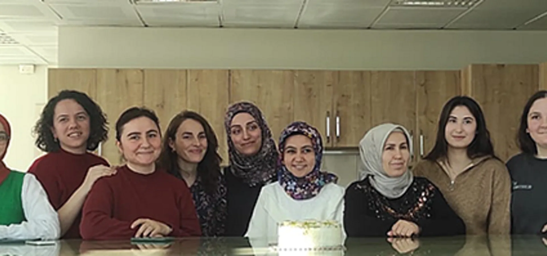 Bir mutfak masasının arkasında yan yana duran on kadın çalışanın toplu fotoğrafı; masada küçük bir pasta duruyor.